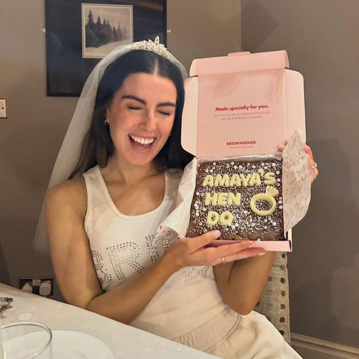 Bride-to-be holding personalised brownie slab reading ‘Amaya’s Hen Do’ with white-chocolate ring icon – delivered to London.