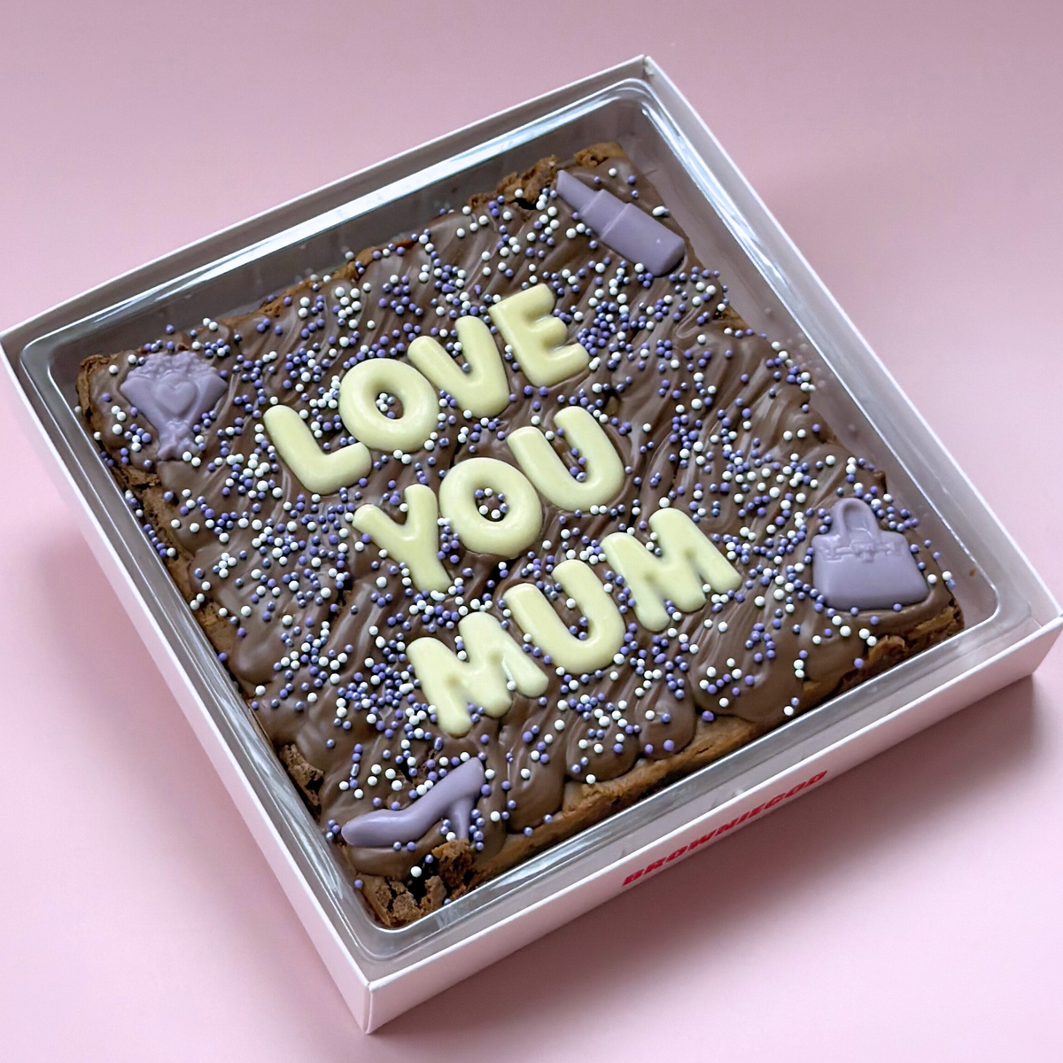 Chocolate brownie slab reading "Love you Mum" in white chocolate letters, with lilac sprinkles and handbag and shoe toppers