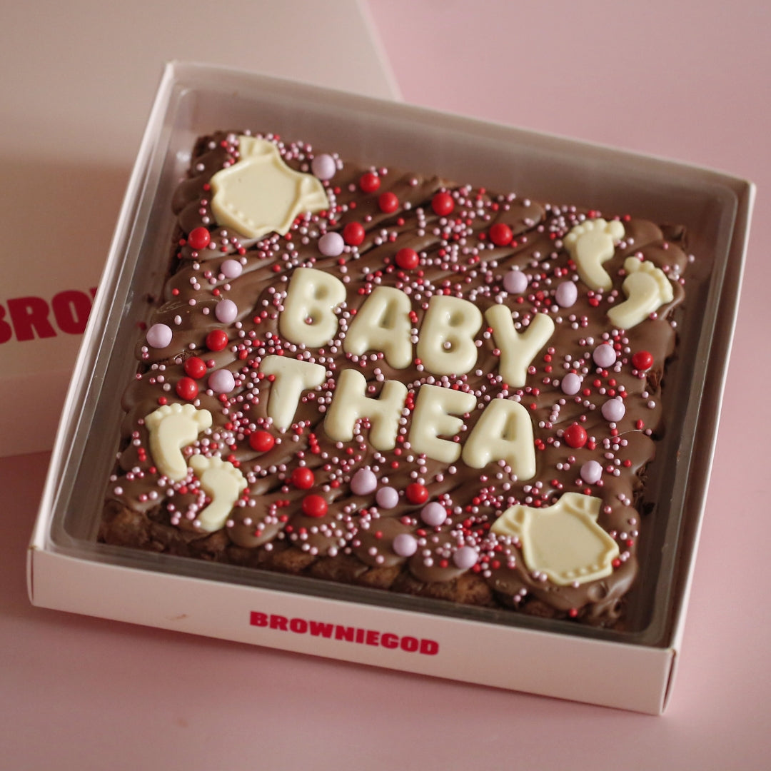Baby brownie slab reading "Baby Thea" in white chocolate letters with onesie and footprint toppers and pink and red sprinkles