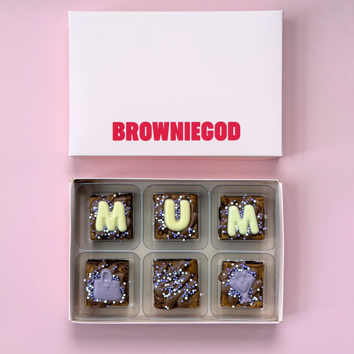Box of six mini brownies spelling "Mum" in white chocolate letters with lilac handbag toppers, next to the white Browniegod box lid