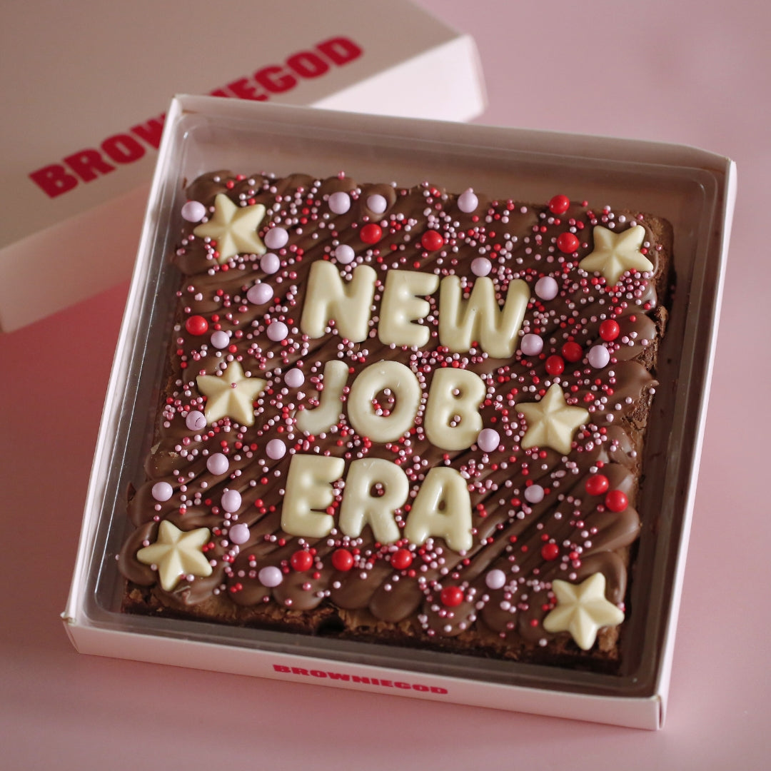 Brownie slab reading "New job era" in white chocolate letters with white star toppers and pink and red sprinkles