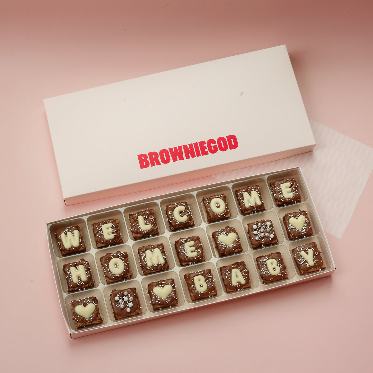 Box of 21 mini brownies spelling "Welcome home baby" in white chocolate letters with heart toppers, next to the box lid