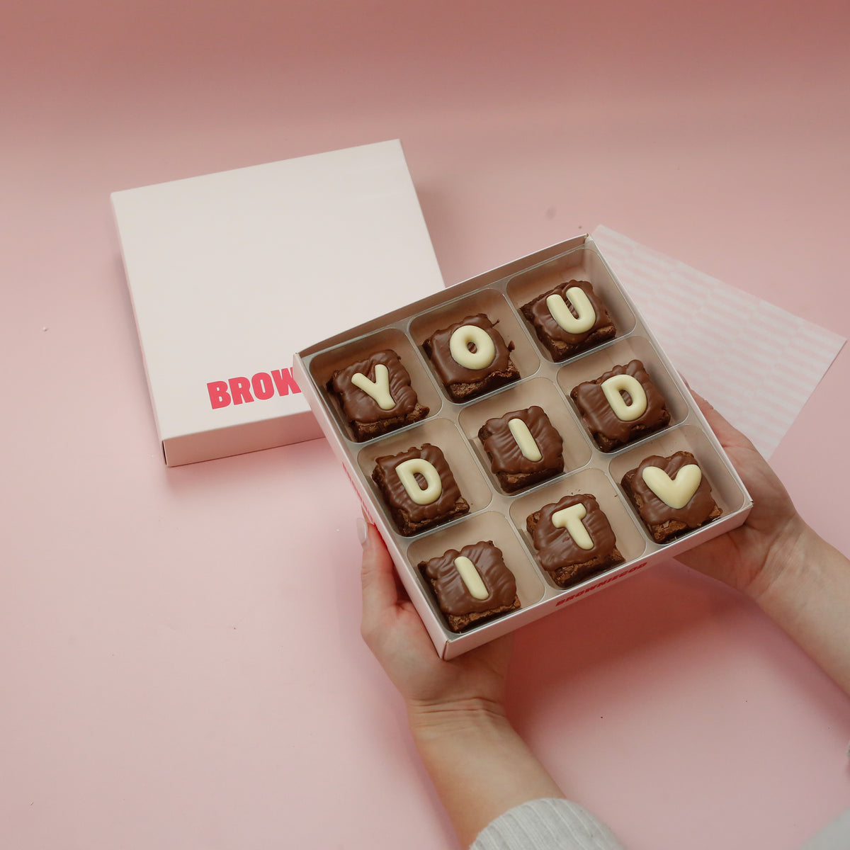 Hands holding a box of nine mini brownies spelling "You did it" in white chocolate letters with a heart topper