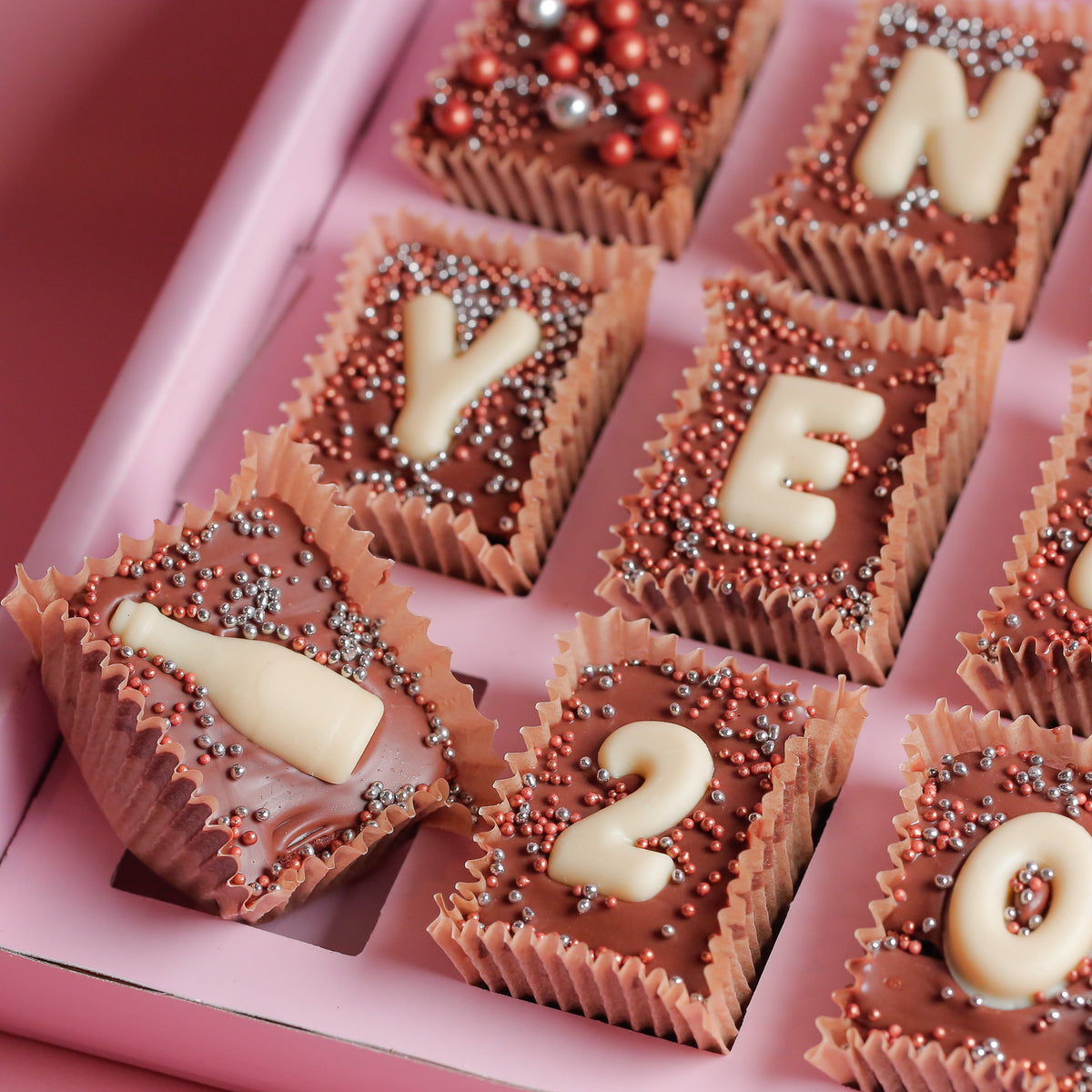 Overhead of New Year mini brownies in pink paper cases, topped with white chocolate letters and numbers and red and pink sprinkles