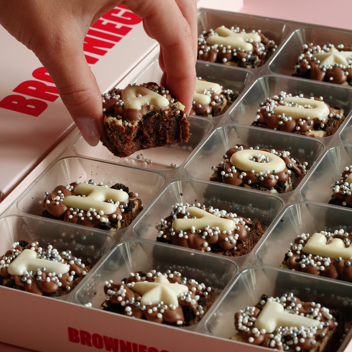 Close-up of a hand holding a bitten mini brownie above a box of white chocolate lettered mini brownies with star toppers