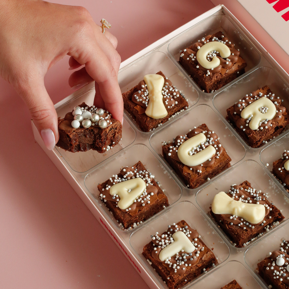 Hand placing a bitten mini brownie into a box of graduation mini brownies with white chocolate letters and scroll toppers