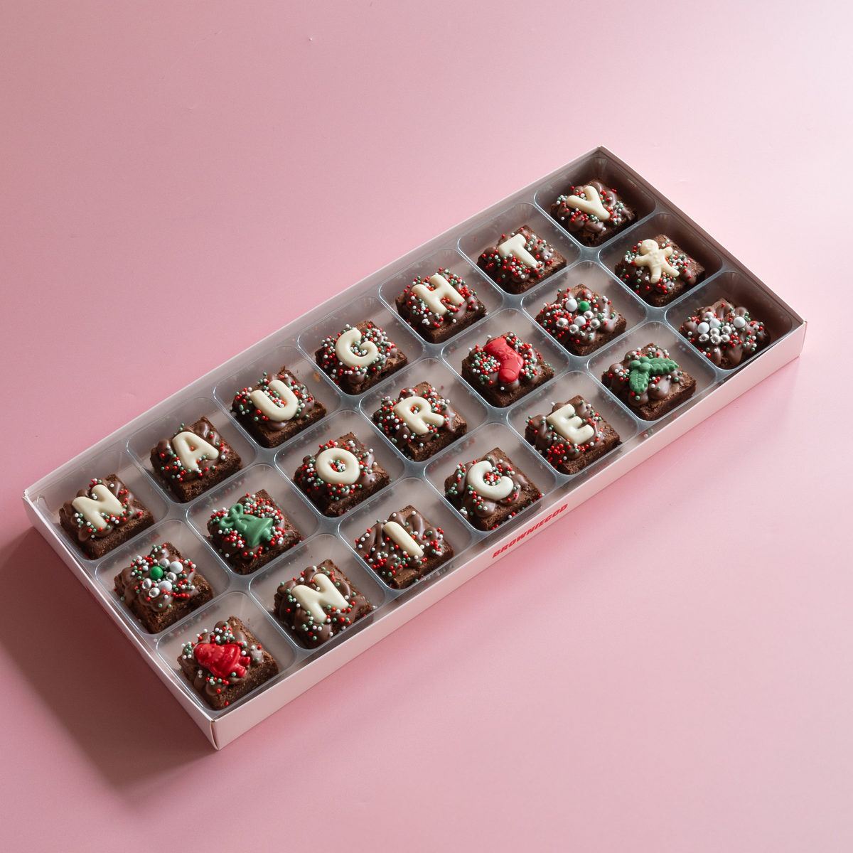 Box of mini brownies spelling "Naughty or nice" in white chocolate letters with red and green Christmas toppers