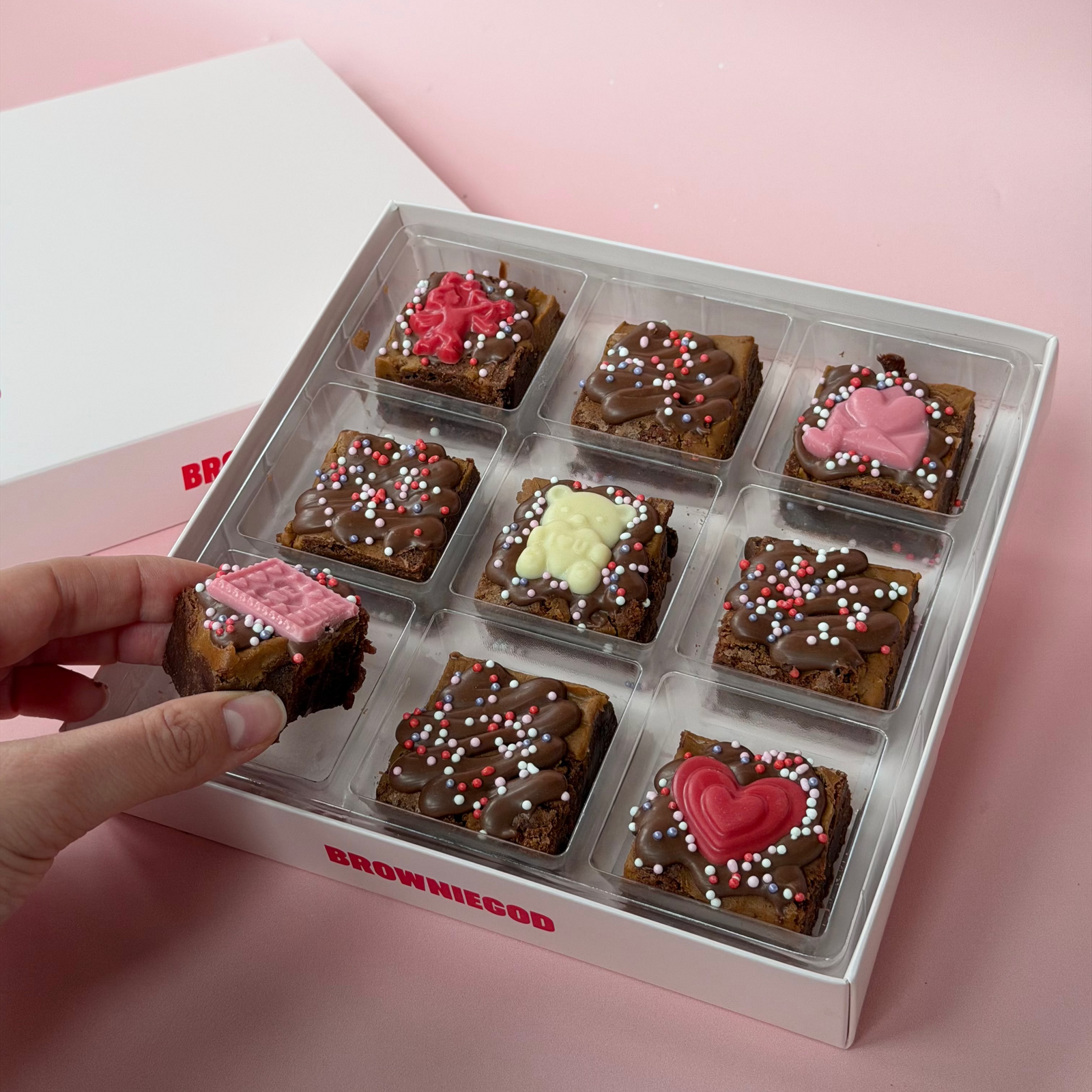 Box of brownies with valentines day decorative toppings held by a hand on a pink background