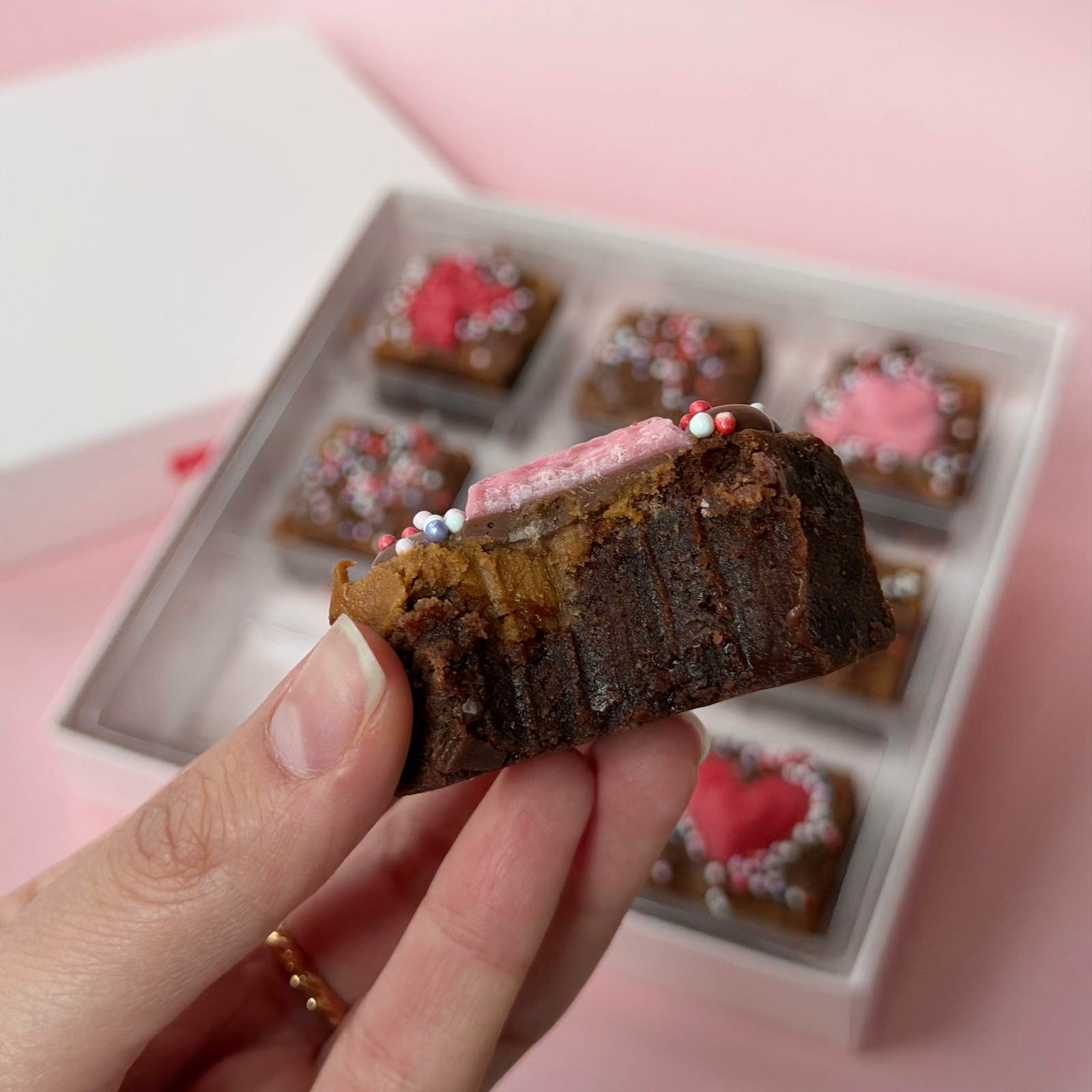 Hand holding a valentines day chocolate brownie with a box of similar desserts in the background on a pink surface