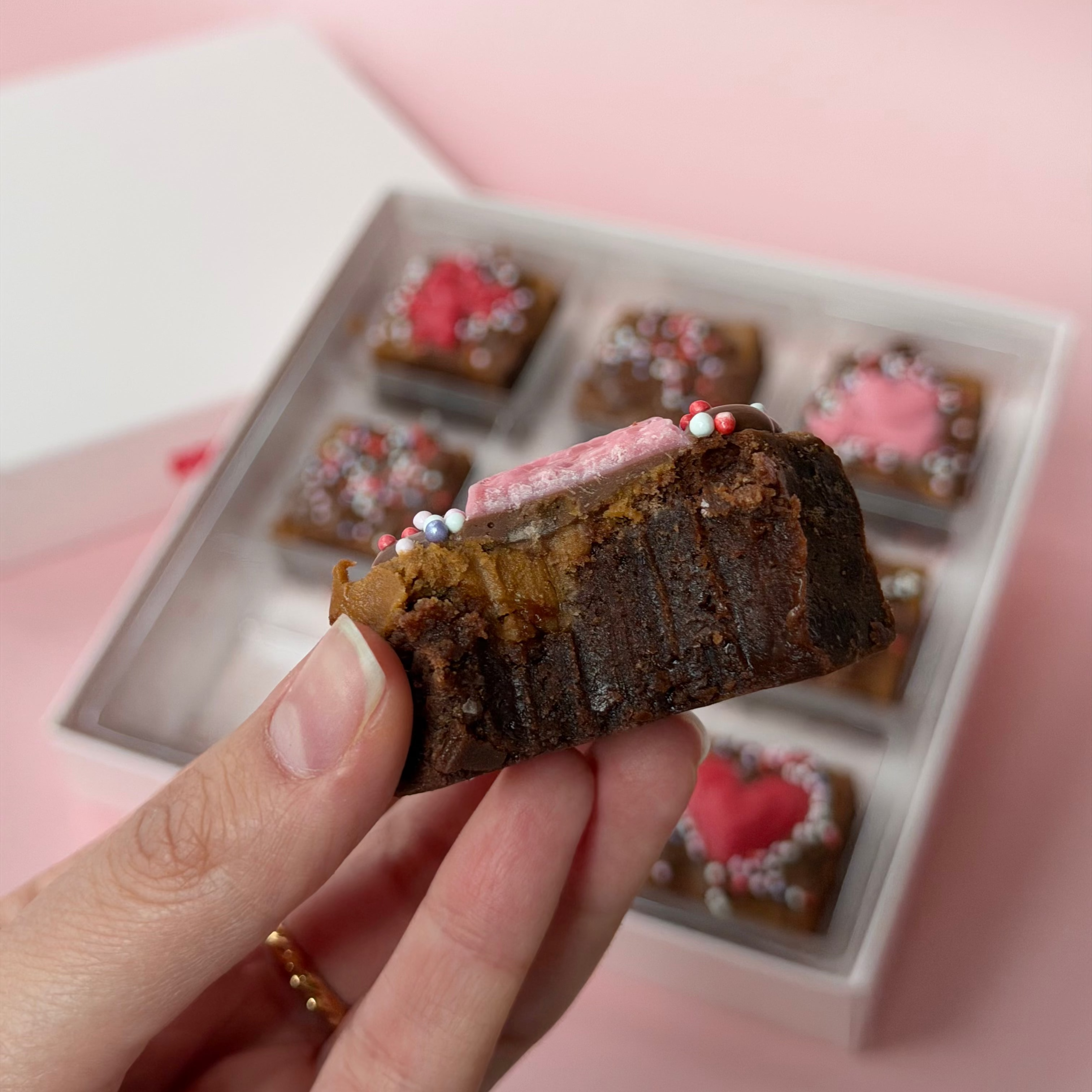 Hand holding a valentines day chocolate brownie with a box of similar desserts in the background on a pink surface