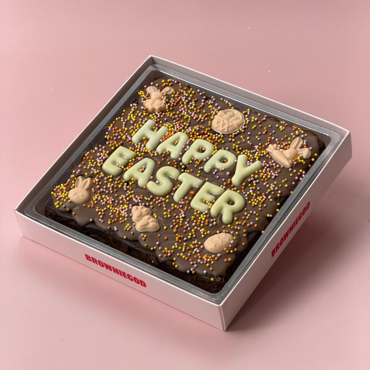 Chocolate brownie slab topped with white chocolate letters reading "Happy Easter", pastel sprinkles and chocolate bunny and egg decorations