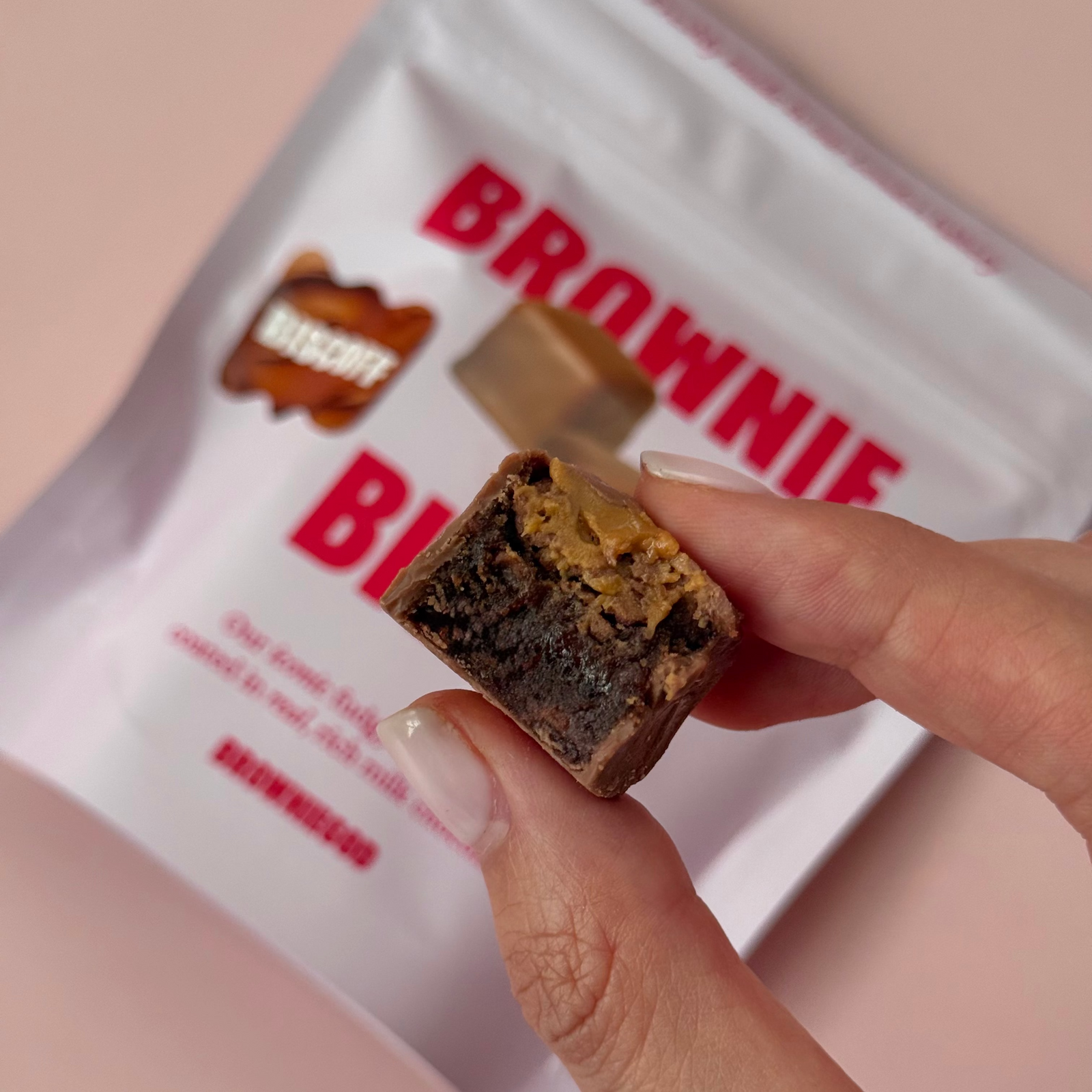Hand holding a bitten Biscoff brownie bite with a layer of Biscoff spread, above a Brownie Bites pouch