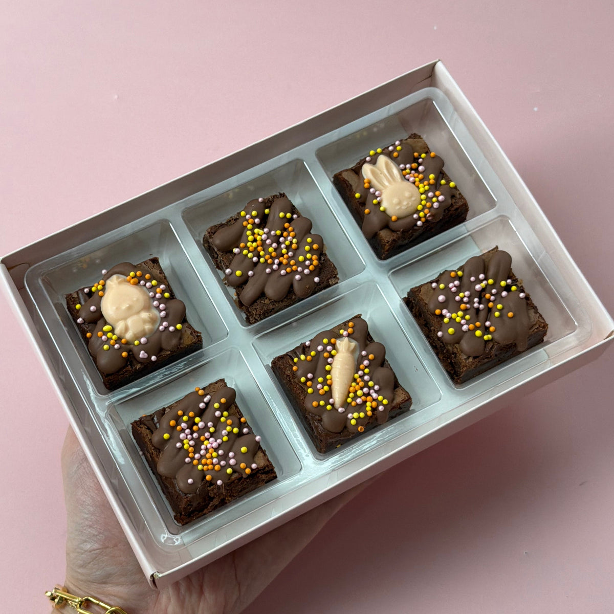 Hand holding a box of six Easter mini brownies topped with chocolate bunnies, carrots and orange and yellow sprinkles