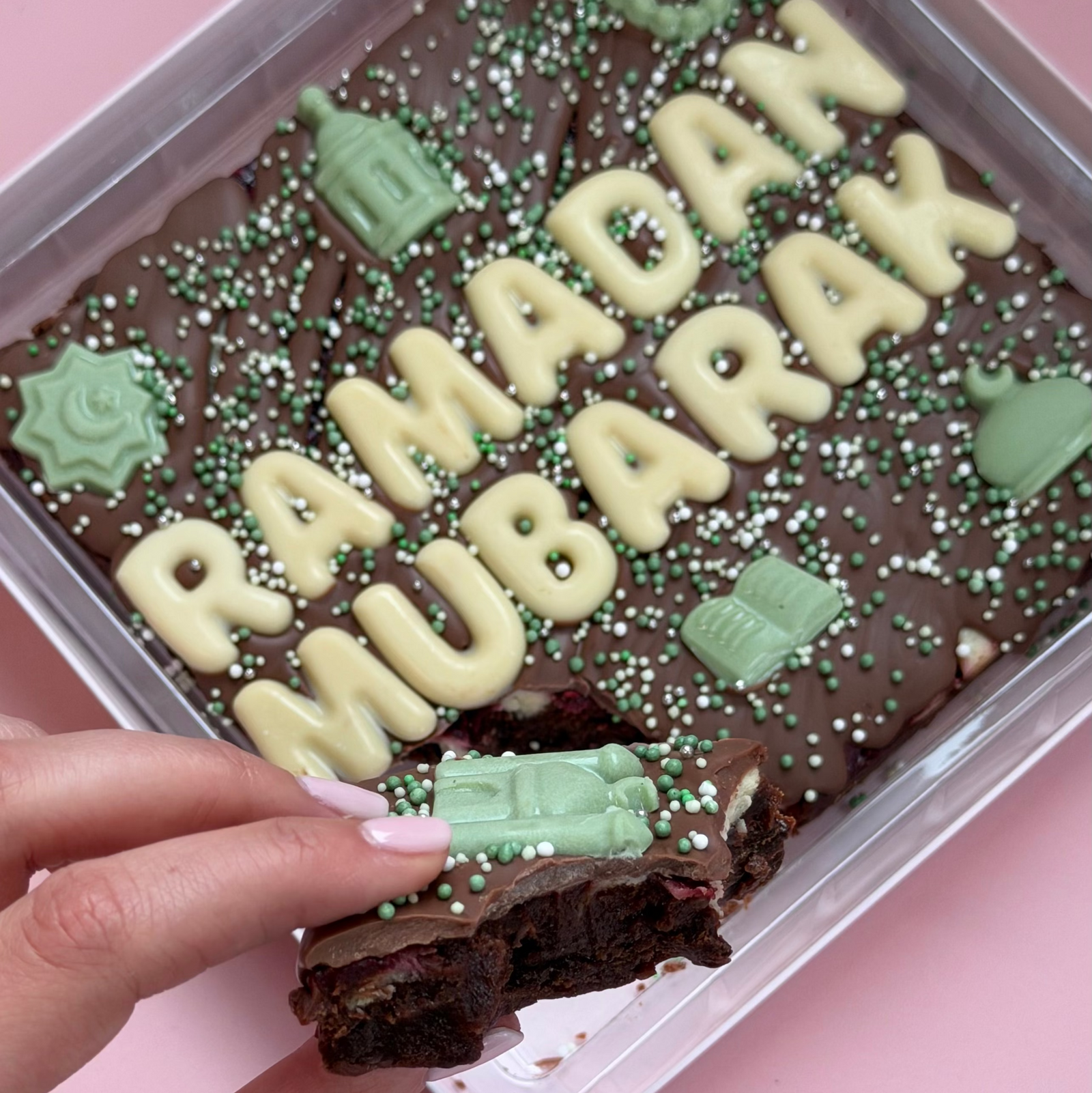 Hand pulling a piece from a "Ramadan Mubarak" brownie slab, showing the fudgy brownie under the chocolate topping